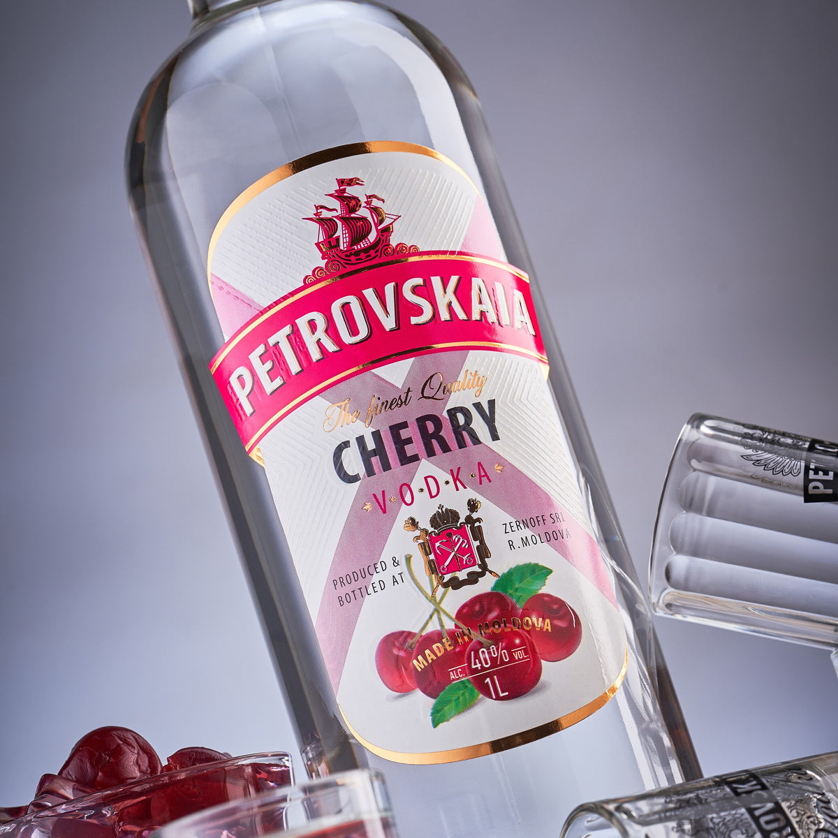 Schuin gefotografeerde 1 liter fles Petrovskaia Cherry Premium Vodka met kersen en shotglaasjes, tegen een grijze achtergrond