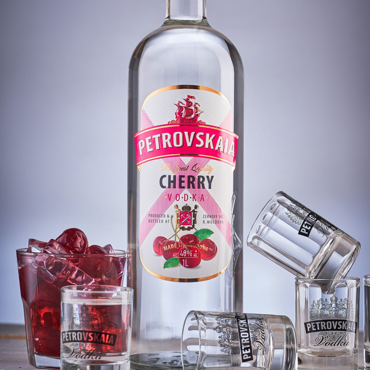 Petrovskaia Cherry Premium Vodka 1 Liter Fles met verse kersen en kersencocktail, omringd door shotglaasjes op een grijze achtergrond