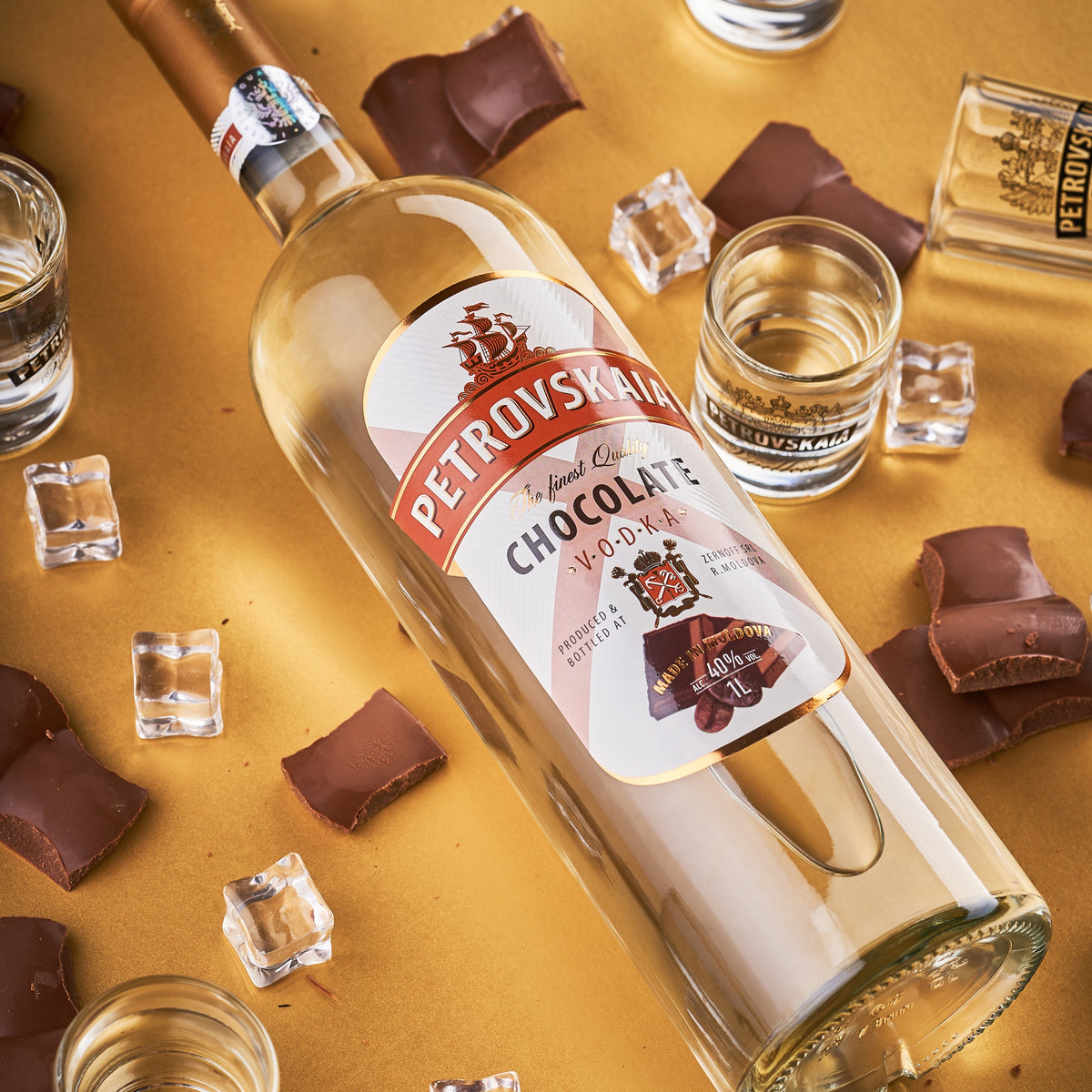 Petrovskaia Chocolate Vodka 1 liter fles schuin liggend tussen stukjes chocolade en ijsblokjes op een gouden achtergrond, luxe drank.